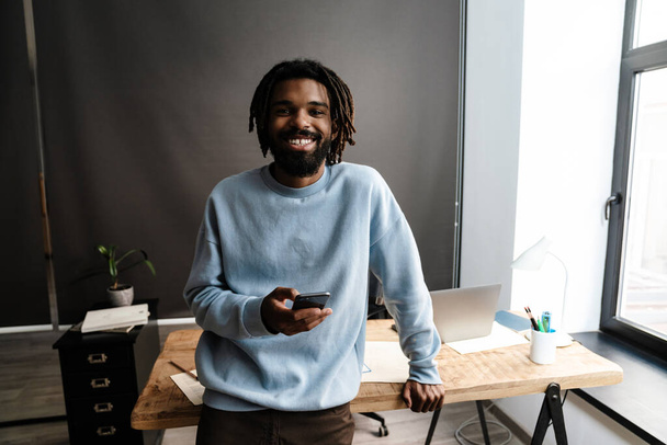 Smiling young african man holding mobile phone standing at the desk - Foto, Imagen