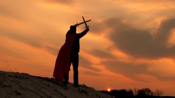 Ο ελεύθερος άνθρωπος παίζει τον ήρωα. Ελεύθερος ιππότης διέσχισε τα σπαθιά του στον ουρανό πάνω από το κεφάλι του. Σούπερμαν με ένα σπαθί στο χέρι του και σε κόκκινο μανδύα στέκεται στο βουνό στο φως ηλιοβασίλεμα. Παιχνίδι της Ρωμαϊκής λεγεώνας. - Πλάνα, βίντεο