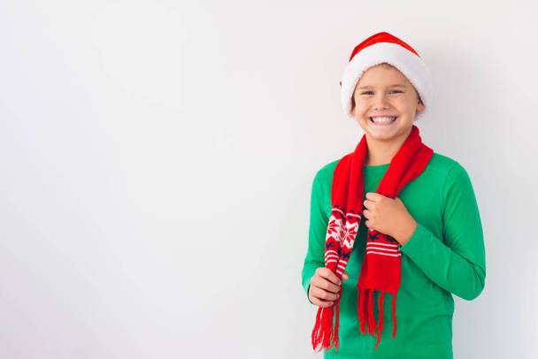 Retrato de niño feliz con sombrero de Santa Claus y bufanda de invierno sobre fondo blanco. Niño divirtiéndose en Navidad. Feliz Navidad concepto - Foto, imagen