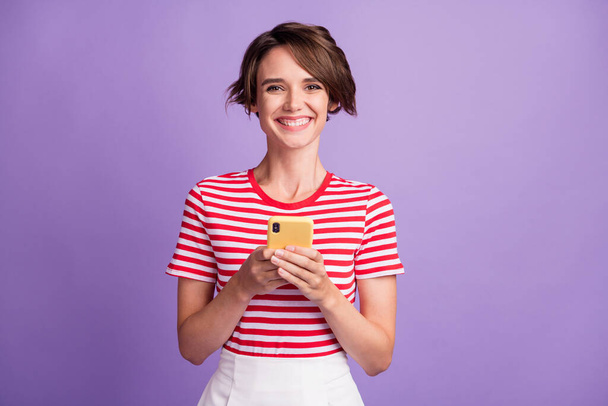 Photo of cheerful pretty lady hold telephone look camera white smile wear striped t-shirt isolated purple color background - Fotografie, Obrázek