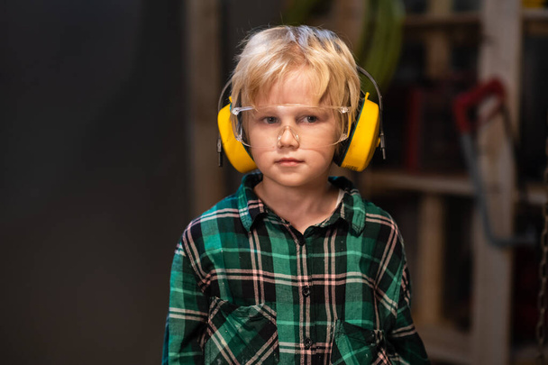 χαριτωμένο αγόρι με ακουστικά και γυαλιά ασφαλείας σε ένα εργαστήριο ξυλουργών, πορτρέτο. - Φωτογραφία, εικόνα