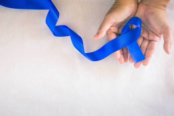 Hands holding deep blue ribbon on white fabric with copy space. Colorectal Cancer Awareness, Colon cancer of older person and world diabetes day, Child abuse prevention. Healthcare, insurance concept. - Photo, image
