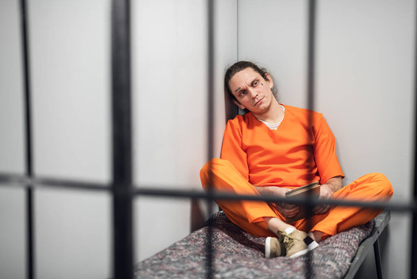 Ένας επικίνδυνος εγκληματίας με τατουάζ στο πρόσωπό του διαβάζει ένα βιβλίο ποίησης που κάθεται σε ένα κελί πίσω από τα κάγκελα. Πήρε στο δρόμο της διόρθωσης. - Φωτογραφία, εικόνα