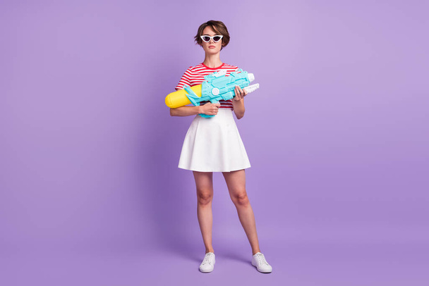 Full length photo of girl hold pump pistol cool look camera wear striped t-shirt sunglass skirt footwear isolated violet color background - Fotó, kép
