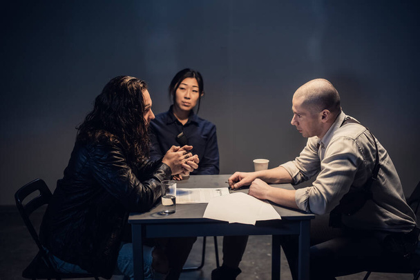 Un detective de policía en la estación y su pareja, una mujer asiática, presentan pruebas a un sospechoso masculino en una sala de entrevistas. - Foto, Imagen