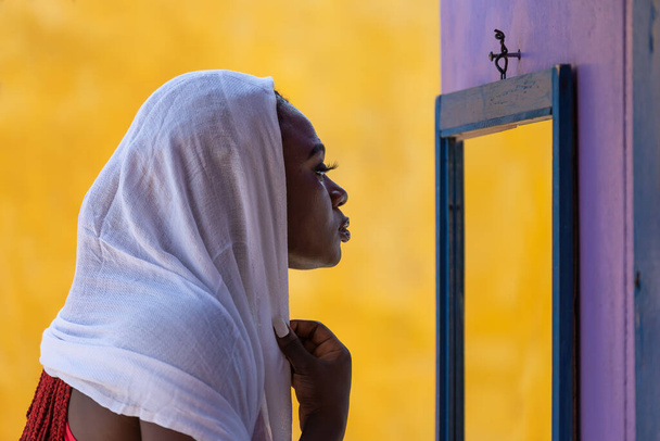 Afrikalı Gana 'lı kadın aynanın önünde beyaz bir şal ile saçını kapatıyor. - Fotoğraf, Görsel
