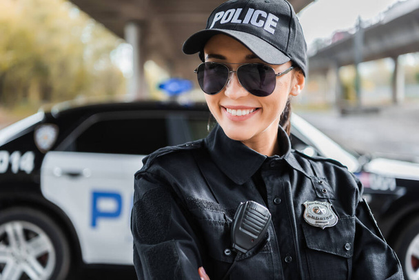 gelukkig jong politievrouw kijken naar camera met wazig patrouille auto op de achtergrond buiten - Foto, afbeelding