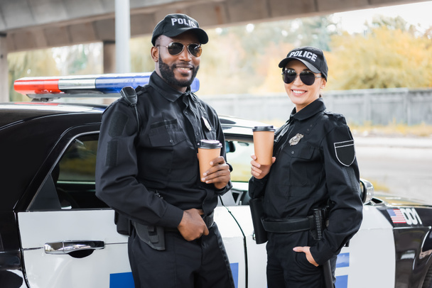 šťastný multikulturní policisté s papírovými poháry při pohledu na kameru v blízkosti hlídkového vozu na rozmazaném pozadí na městské ulici - Fotografie, Obrázek