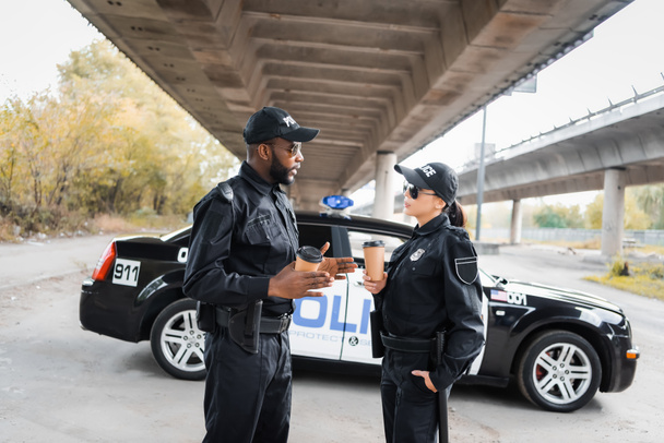 Kağıt bardaklı çok kültürlü polis memurları şehir sokaklarının bulanık arka planında devriye arabasının yanında konuşuyorlar. - Fotoğraf, Görsel