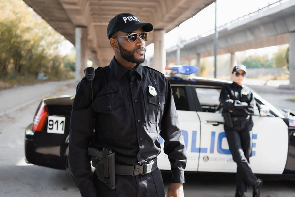 ernstige Afro-Amerikaanse politieman weg te kijken met wazig collega en patrouille auto op de achtergrond op stedelijke straat - Foto, afbeelding
