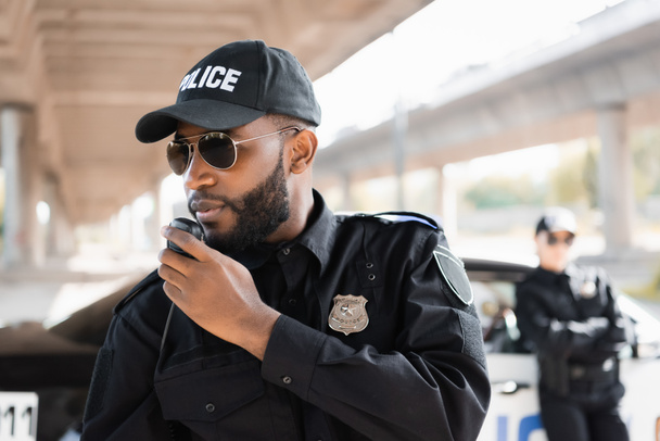 Αφροαμερικάνος αστυνομικός μιλάει στο ραδιόφωνο κοντά σε αστυνομικίνα σε θολή φόντο σε εξωτερικούς χώρους - Φωτογραφία, εικόνα