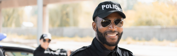 portret van gelukkig Afrikaans amerikaanse politieagent op zoek naar camera met wazig collega op de achtergrond buiten, banner - Foto, afbeelding