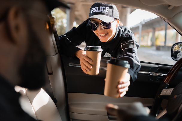 happy policewoman giving paper cup to african american colleague in patrol car on blurred foreground - Fotoğraf, Görsel