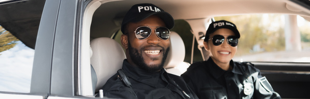 oficiales de policía multiculturales felices mirando la cámara en el coche patrulla sobre fondo borroso, pancarta - Foto, Imagen