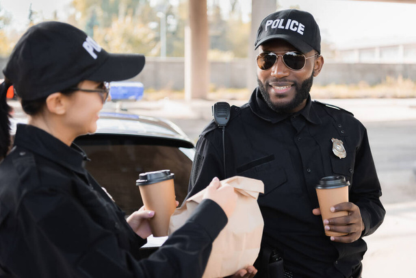 gelukkig politievrouw met papier beker geven pakket naar Afrikaans amerikaanse collega in de buurt patrouille auto op wazig achtergrond buiten - Foto, afbeelding