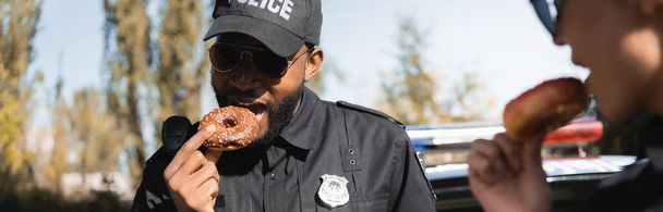 Afrikaans Amerikaans politieagent eet donut met wazig collega op de voorgrond buiten, banner - Foto, afbeelding