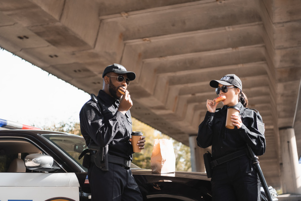 multicultural police officers with paper cups eating doughnuts near patrol car on blurred background on urban street - Foto, Imagen