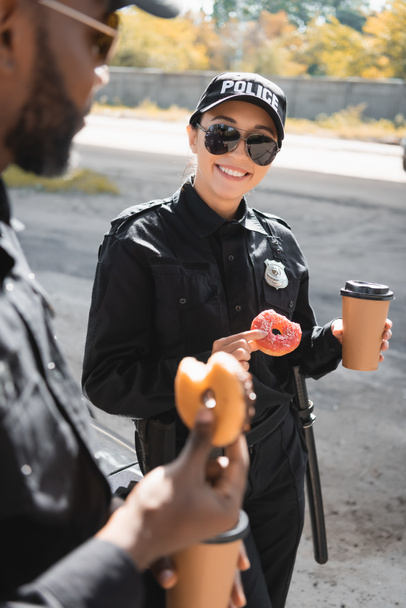 Ευτυχισμένη αστυνομικίνα με ντόνατ και χάρτινο κύπελλο να στέκεται κοντά σε Αφροαμερικανό συνάδελφο σε θολή πρόσοψη σε εξωτερικούς χώρους - Φωτογραφία, εικόνα