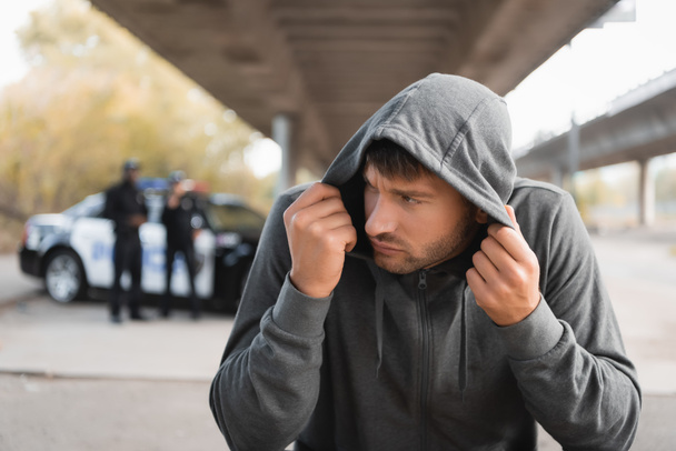 frightened hooded offender hiding with blurred police officers on background outdoors - Photo, Image