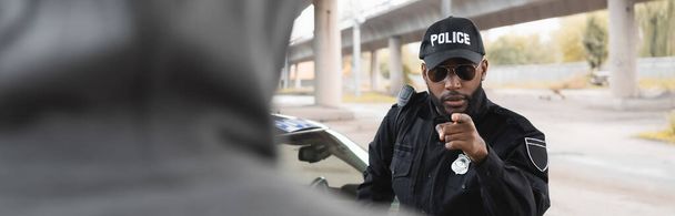 policía afroamericana seria señalando con el dedo al delincuente encapuchado borroso en primer plano en la calle urbana, pancarta - Foto, imagen