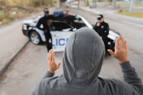 vista trasera del delincuente encapuchado mostrando las manos con oficiales de policía multiculturales borrosas en el fondo al aire libre - Foto, imagen