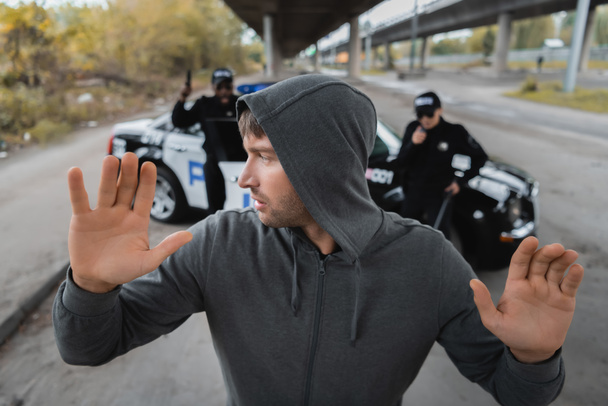 Elleri havada kapüşonlu suçlu, şehir sokaklarında arka planda bulanık, çok kültürlü polis memurlarına bakıyor. - Fotoğraf, Görsel