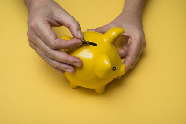 Closeup of hands of man insert one euro coin in his yellow piggy bank on yellow background - Valokuva, kuva