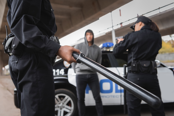 policier afro-américain tenant matraque près de son collègue visant avec pistolet et délinquant sur fond flou à l'extérieur - Photo, image