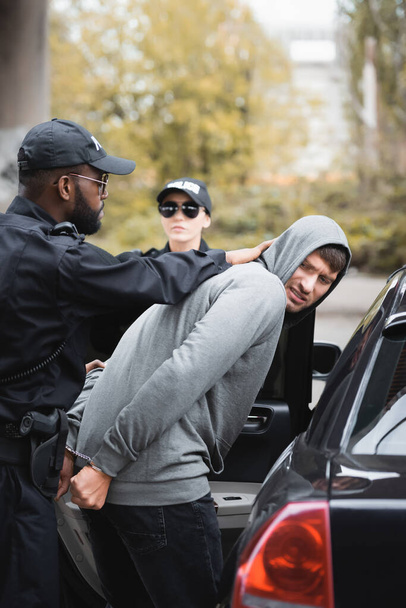Afro-Amerikan polisi, arka planda bulanık bir şekilde meslektaşı ve devriye arabasının yanında öfkeli kapüşonlu suçluyu tutukladı. - Fotoğraf, Görsel