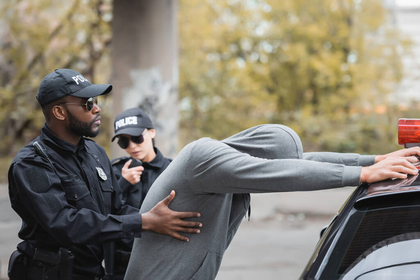 policía afroamericano registrando a delincuente encapuchado apoyado en patrulla cerca de colega sobre fondo borroso al aire libre - Foto, Imagen