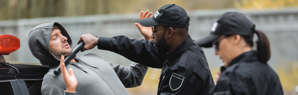 poliziotto afroamericano con manganello che arresta il criminale incappucciato con collega sfocato in primo piano all'aperto, striscione - Foto, immagini