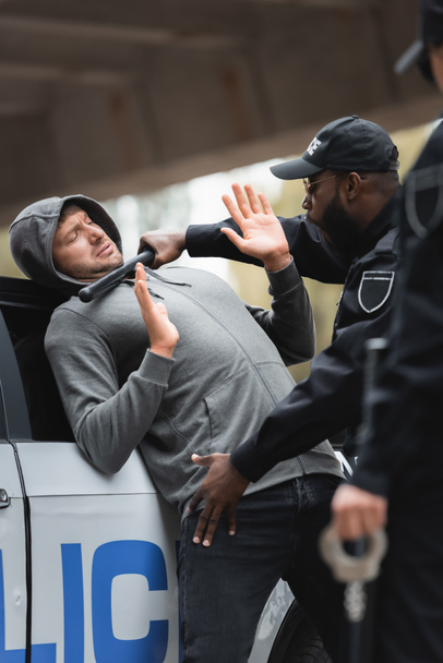 poliziotto afroamericano con truncheon perquisire criminale incappucciato con le mani alzate su primo piano sfocato all'aperto - Foto, immagini