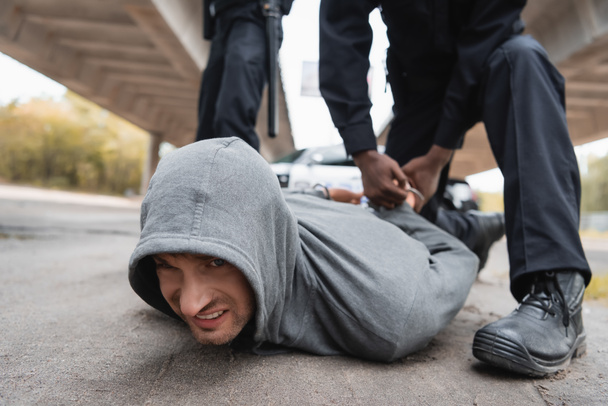 policía afroamericano esposando a delincuente acostado en la calle sobre fondo borroso al aire libre - Foto, imagen
