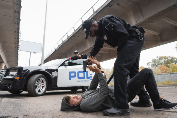 tutta la lunghezza del poliziotto afro-americano mirando con la pistola al criminale spaventato sdraiato sulla strada urbana su sfondo sfocato - Foto, immagini