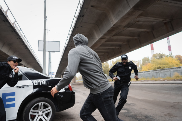 délinquant à capuchon fuyant des policiers multiculturels près d'une voiture de patrouille dans une rue urbaine - Photo, image