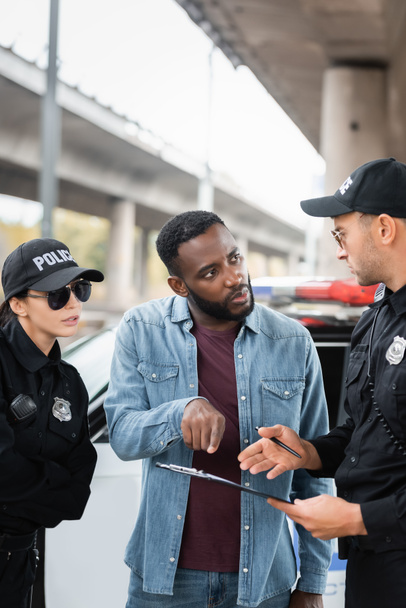 african american victim pointing with finger at clipboard while talking with policeman on blurred background outdoors - Foto, imagen