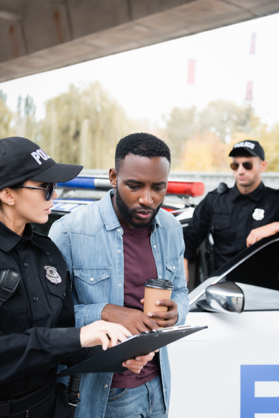 Politievrouw houdt klembord in de buurt van Afrikaans Amerikaans slachtoffer met koffie te gaan en collega in de buurt van auto op wazige achtergrond  - Foto, afbeelding
