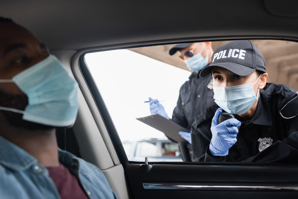 Policjantka w masce medycznej za pomocą krótkofalówki w pobliżu kolegi ze schowkiem i amerykańskim kierowcą na zamazanym pierwszym planie w samochodzie  - Zdjęcie, obraz