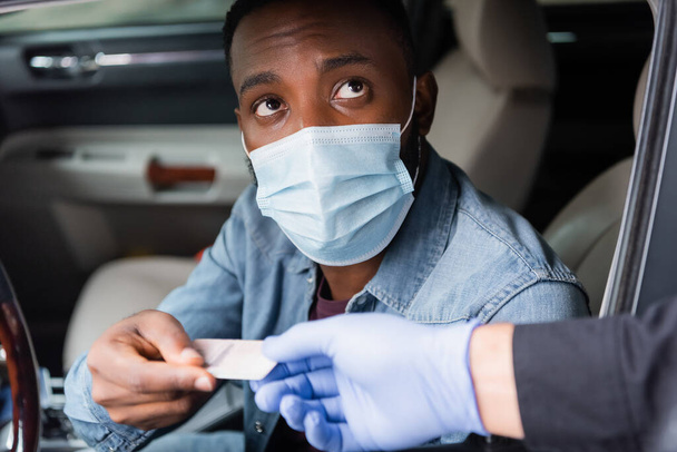 Tıbbi maskeli Afrikalı Amerikalı sürücü arabanın yakınındaki bulanık ön plandaki polise ehliyet veriyor.  - Fotoğraf, Görsel