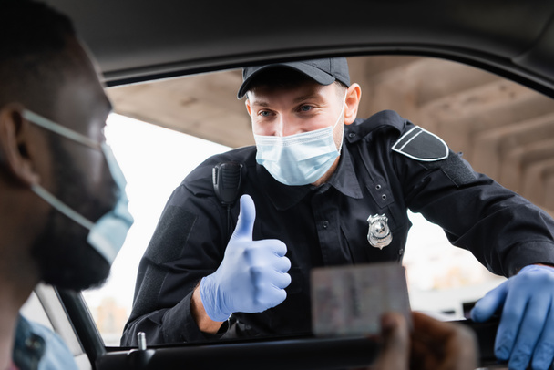 Polizist mit medizinischer Maske und Latex-Handschuhen zeigt afrikanisch-amerikanischen Fahrer mit Führerschein auf verschwommenem Vordergrund im Auto  - Foto, Bild