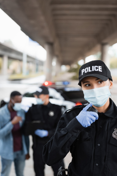 Policewoman en masque médical utilisant talkie walkie près de victime afro-américaine et collègue sur fond flou dans la rue urbaine  - Photo, image