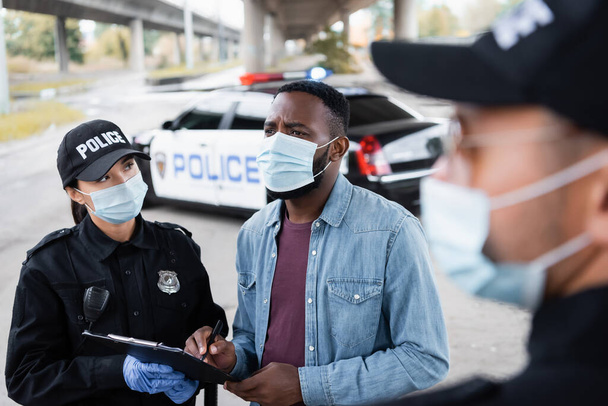 Африканская американская жертва в медицинской маске держит ручку возле офицера полиции с планшетом и полицейским на размытом переднем плане  - Фото, изображение