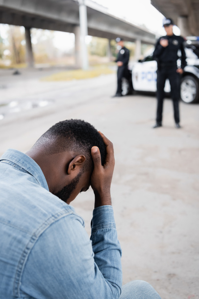 Triste víctima afroamericana cerca de oficiales de policía y coche sobre fondo borroso al aire libre  - Foto, imagen