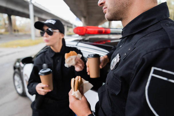 Politieagent met hamburger en koffie te gaan in de buurt van collega en auto op wazige achtergrond op stedelijke straat  - Foto, afbeelding
