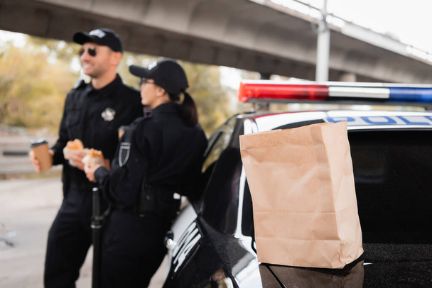 Paper bag on trunk of car near police officers with burgers and coffee to go on blurred background on urban street  - Photo, Image