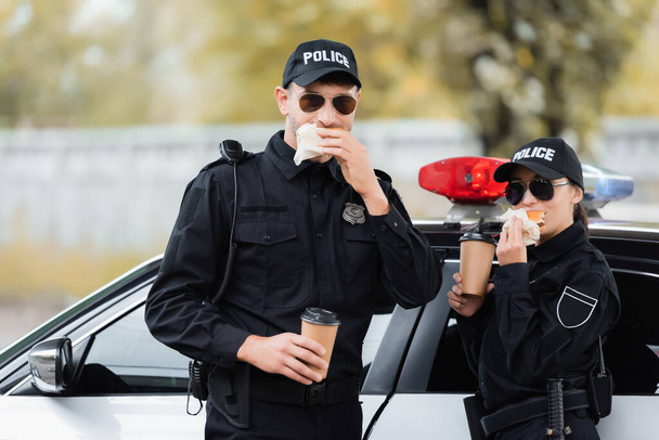 Policías en gafas de sol comiendo hamburguesas y sosteniendo café para llevar cerca del coche al aire libre  - Foto, Imagen