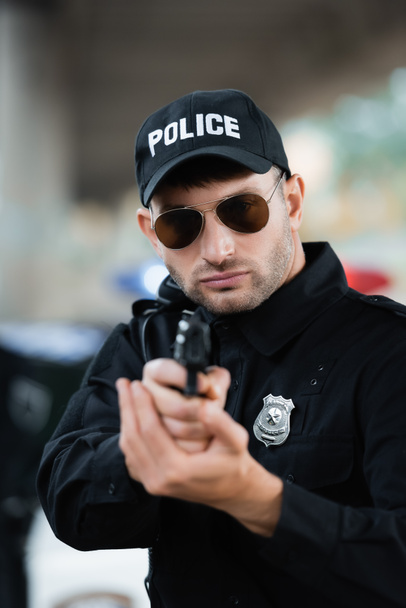 Police officer in uniform and sunglasses holding gun on blurred foreground outdoors  - Foto, afbeelding