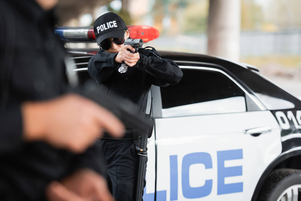 Policewoman in sunglasses holding gun near colleague on blurred foreground and car outdoors  - Photo, Image