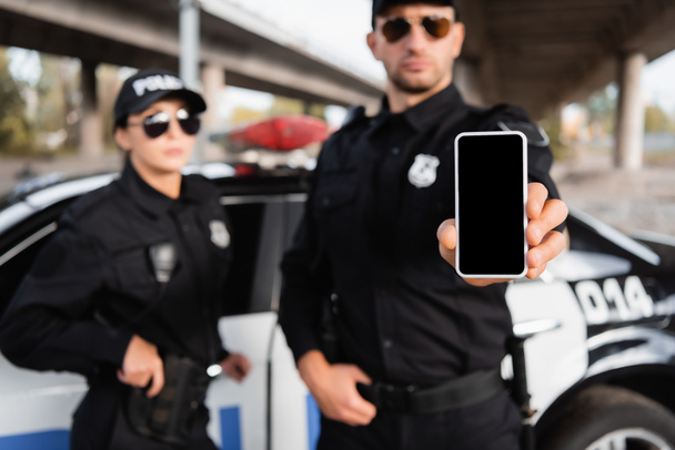 Smartphone mit leerem Bildschirm in der Hand eines Polizisten in der Nähe von Kollege und Auto auf verschwommenem Hintergrund  - Foto, Bild
