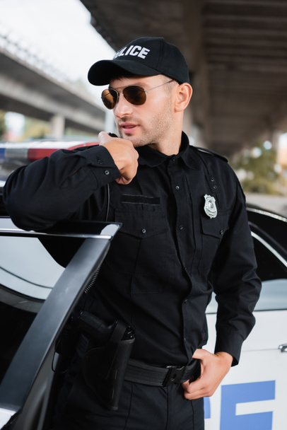 Policeman in sunglasses using walkie talkie near car on urban street  - Фото, изображение